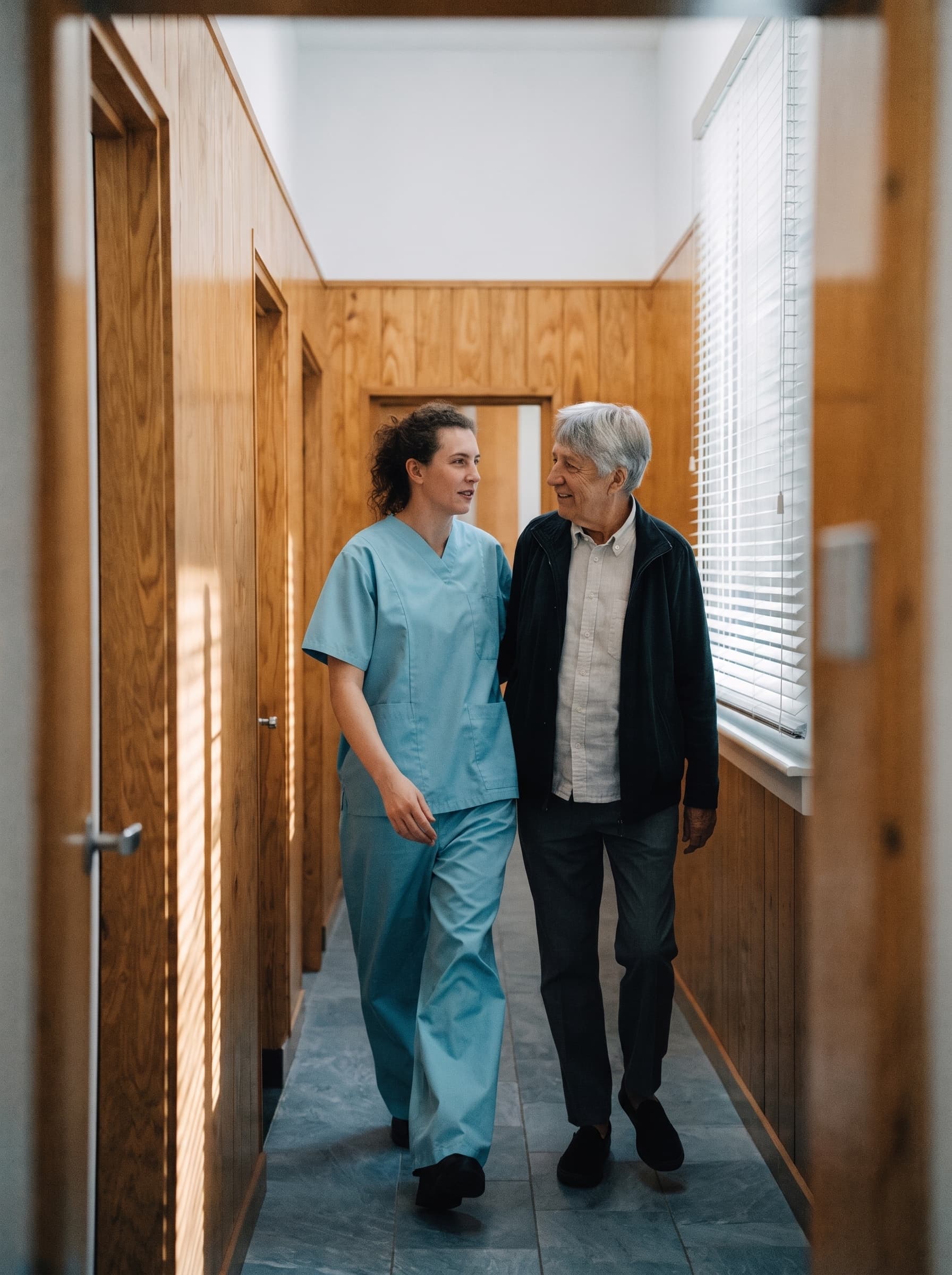 A caregiver in scrubs walking beside an older patient along a sunlit, wood-panelled office corridor