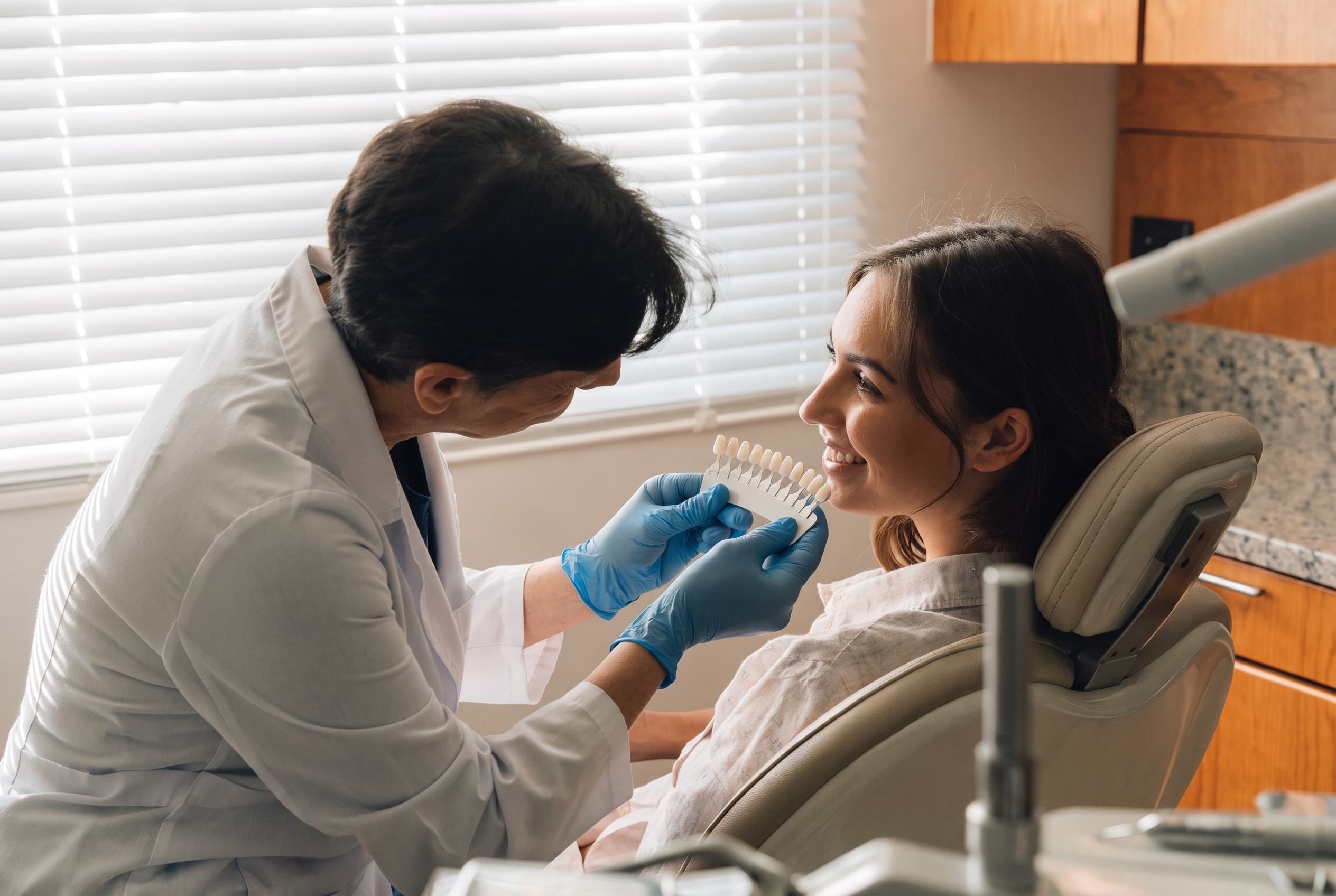 A clinician in a white coat holding a row of tooth shade tabs up beside the smile of a woman in the treatment chair, comparing the colour