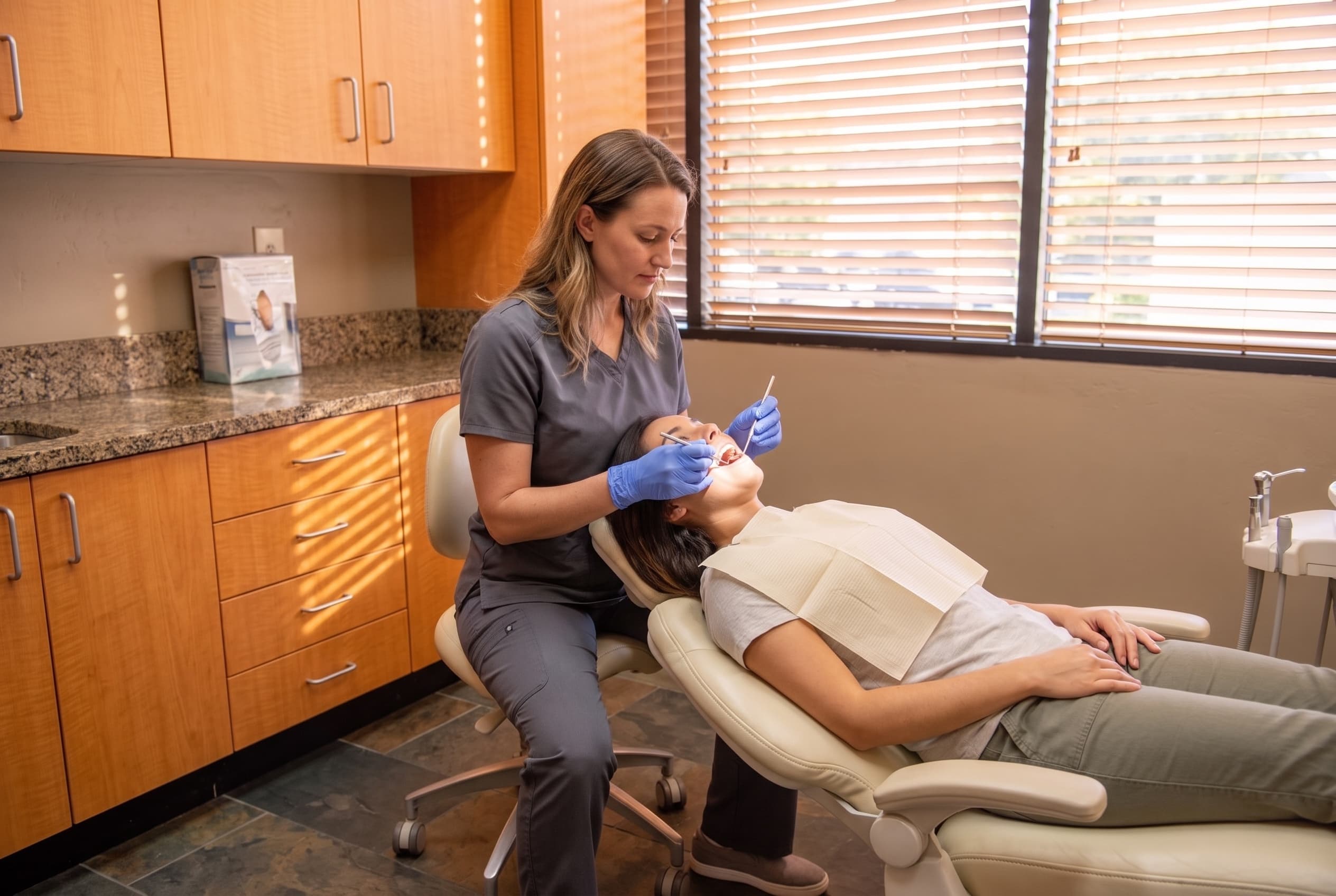 A hygienist working chairside on a reclined patient with a dental mirror and explorer probe