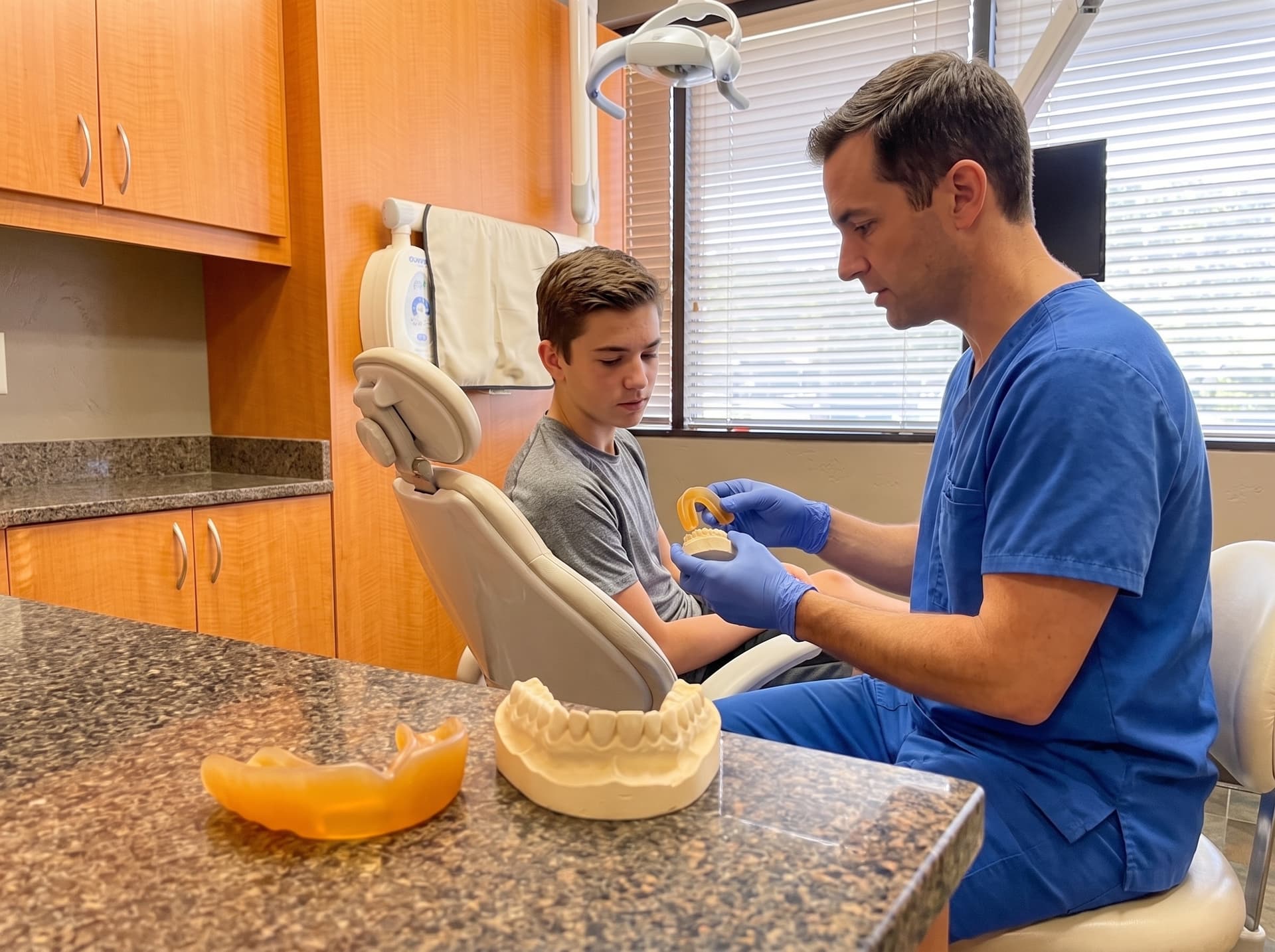 A clinician holding a yellow sports mouthguard fitted on a stone model for a teenage boy in the treatment chair, a second guard and a plaster model on the counter in the foreground