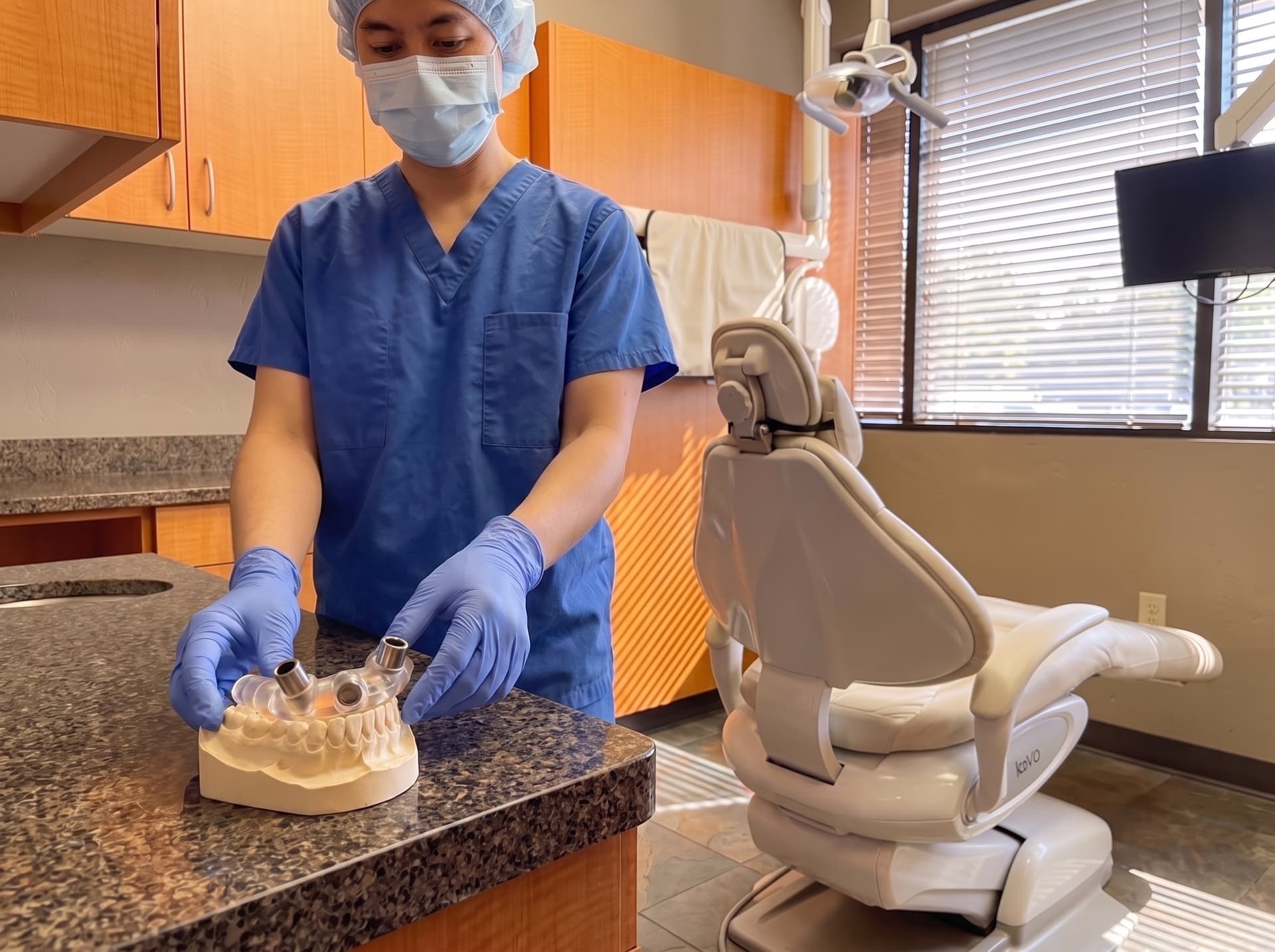 A clinician in surgical cap and mask steadying a clear surgical guide over a stone dental model on the counter, two metal guide sleeves standing up from it