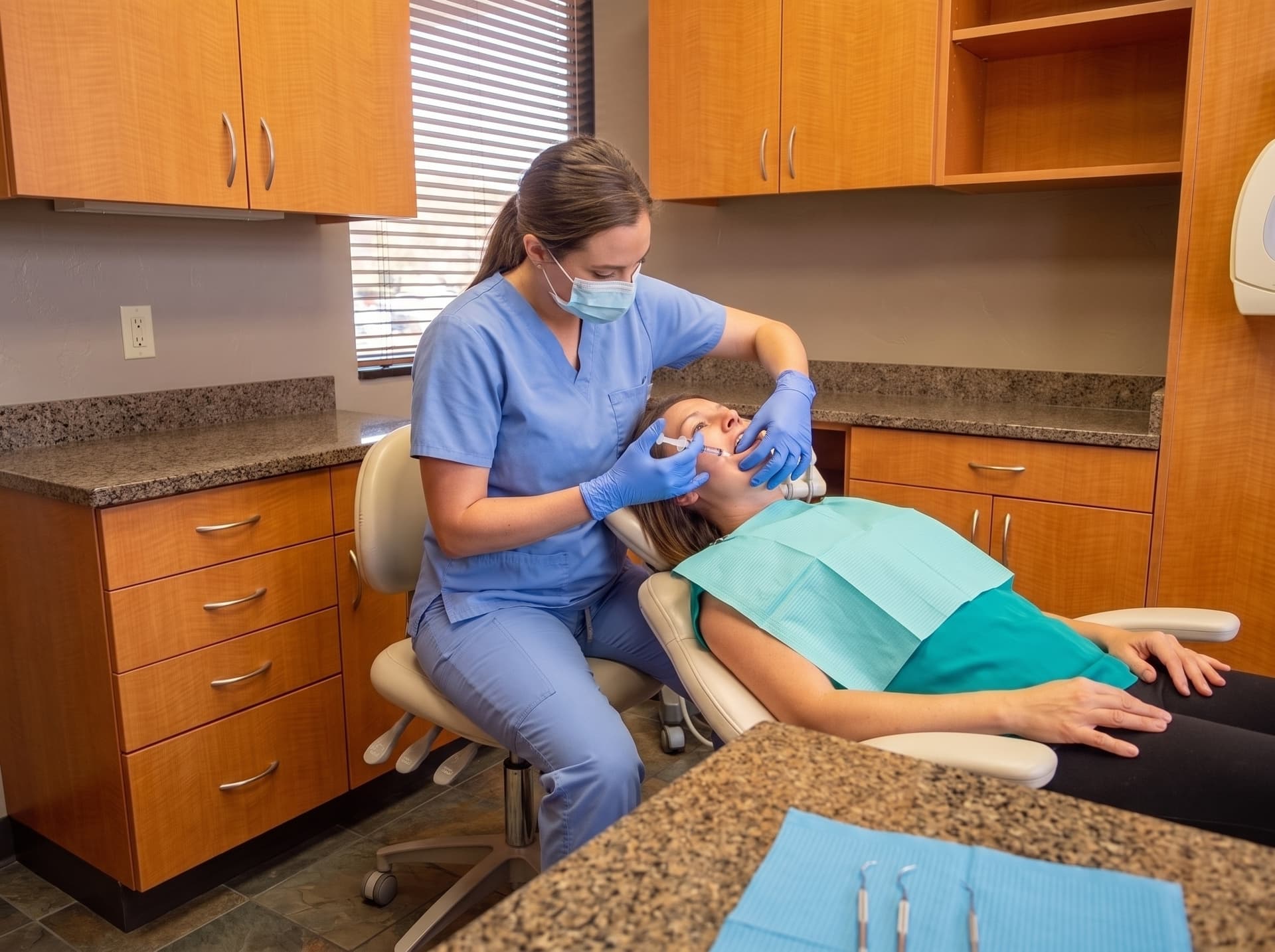 A hygienist in a mask working along the gum line of a reclined patient in a teal bib, periodontal instruments laid out on a blue field in the foreground