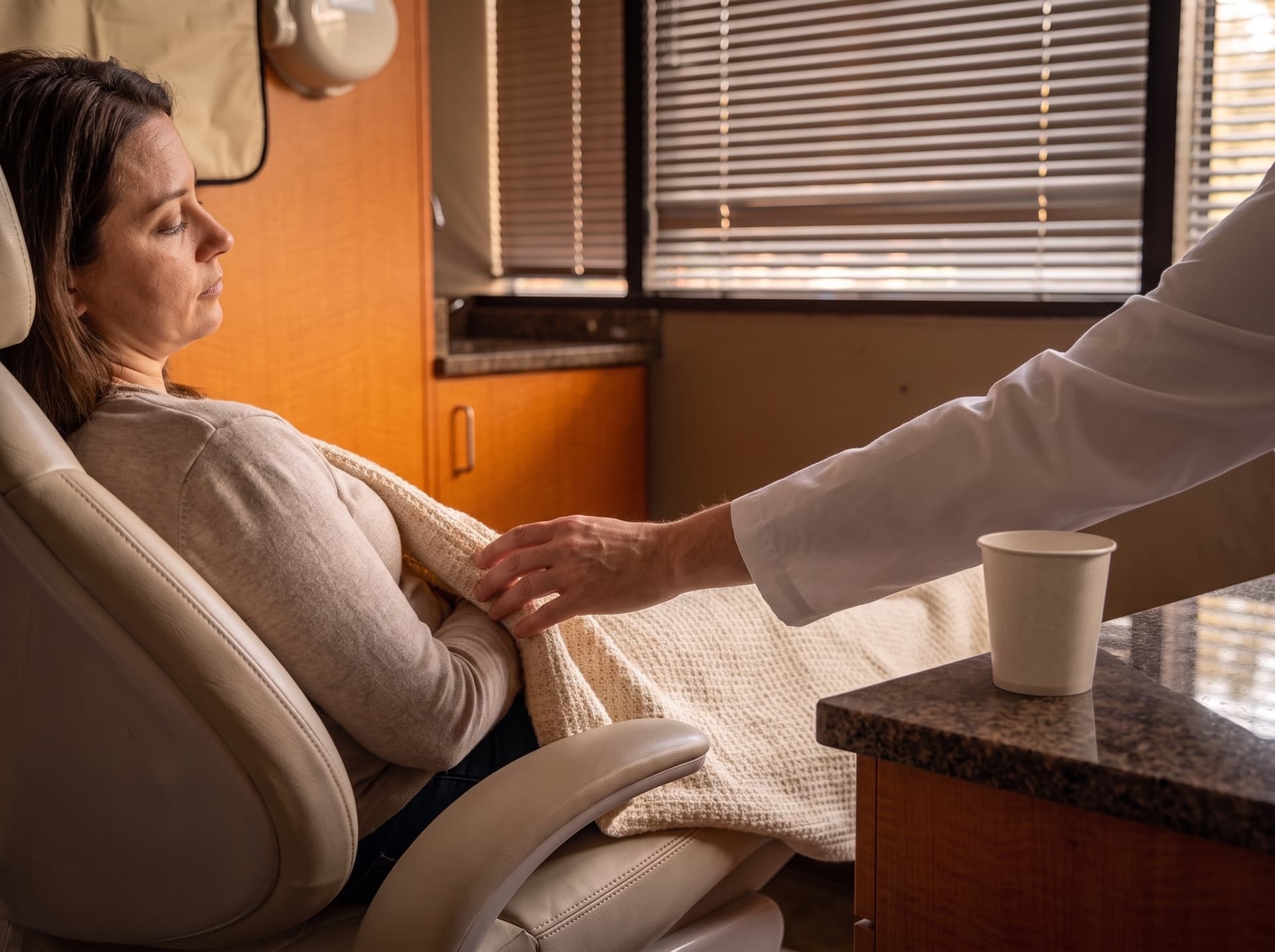 A patient settled in the treatment chair under a knit blanket, a clinician's hand steadying it, a cup of water on the ledge