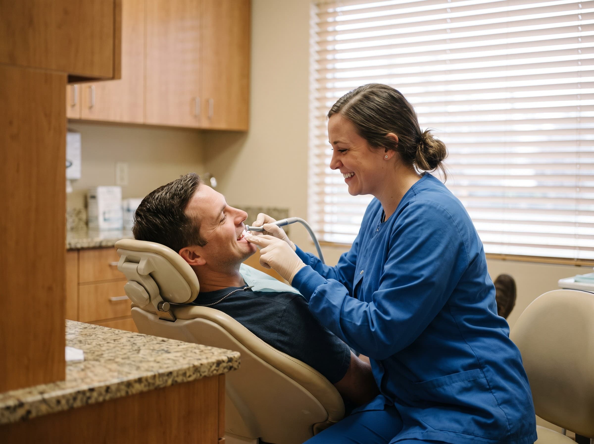 A hygienist cleaning a reclined man's teeth with a handpiece, both of them laughing, a granite counter cutting across the foreground