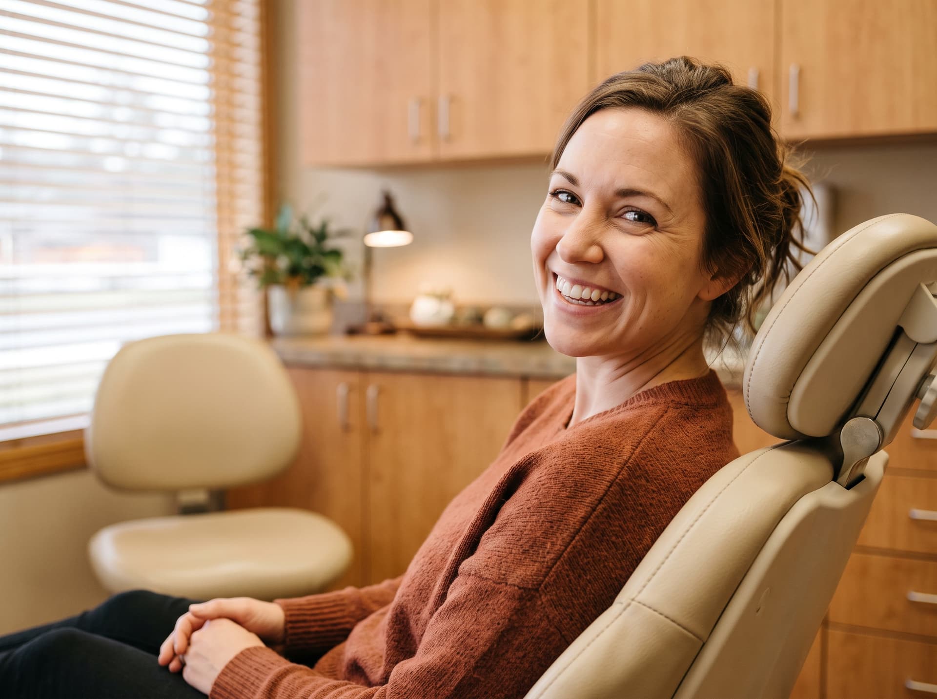 A woman in a rust knit sweater sitting in the treatment chair and laughing toward the camera, warm daylight coming through the blinds behind her