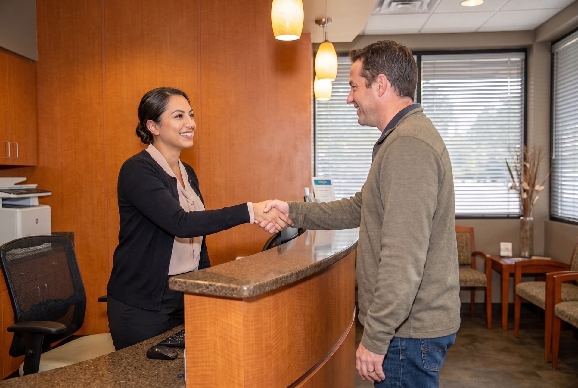 A receptionist behind the maple and granite front desk shaking hands with a patient arriving for an appointment