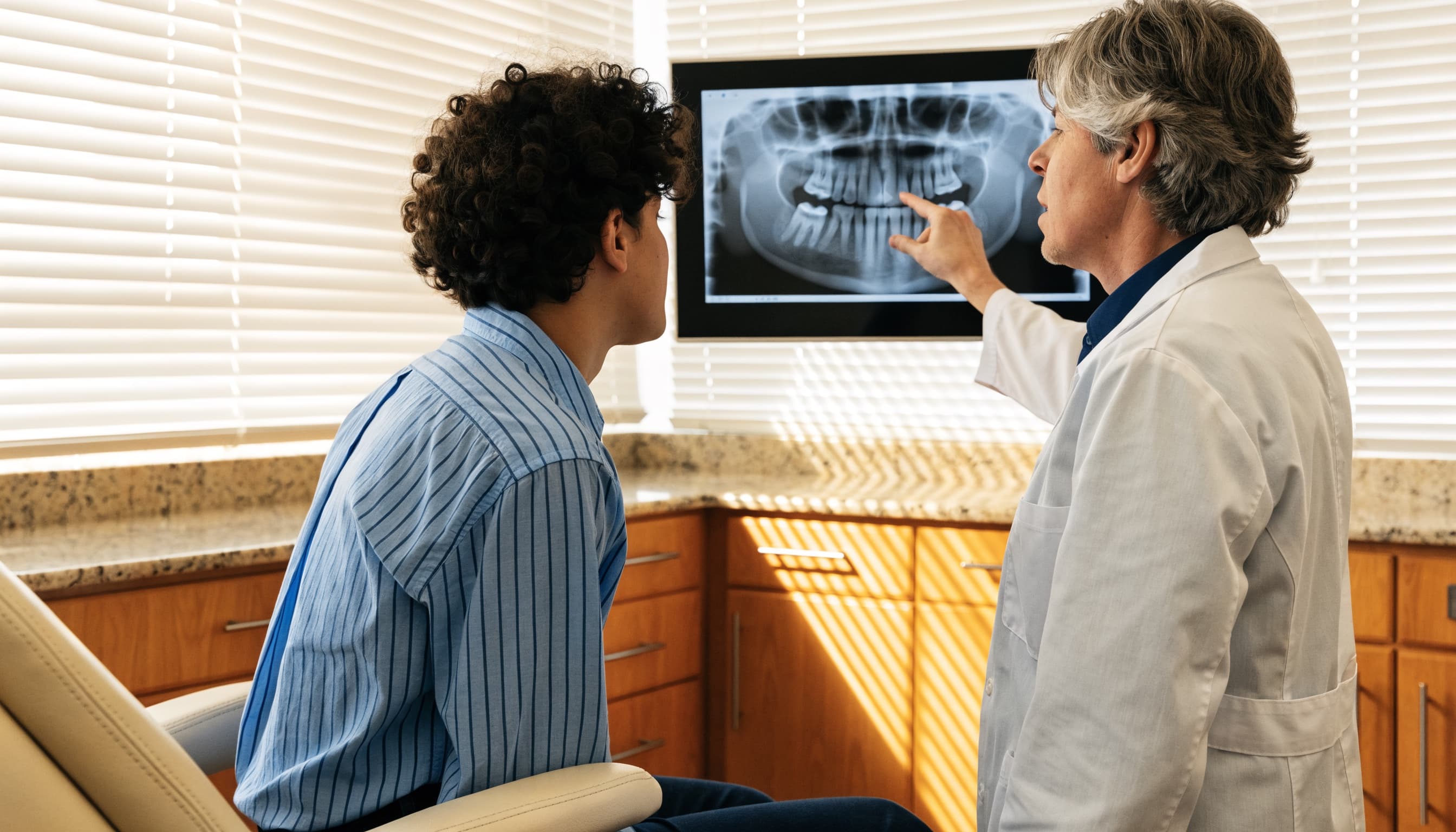 A clinician pointing at a panoramic dental radiograph on a wall screen while a seated patient looks at it
