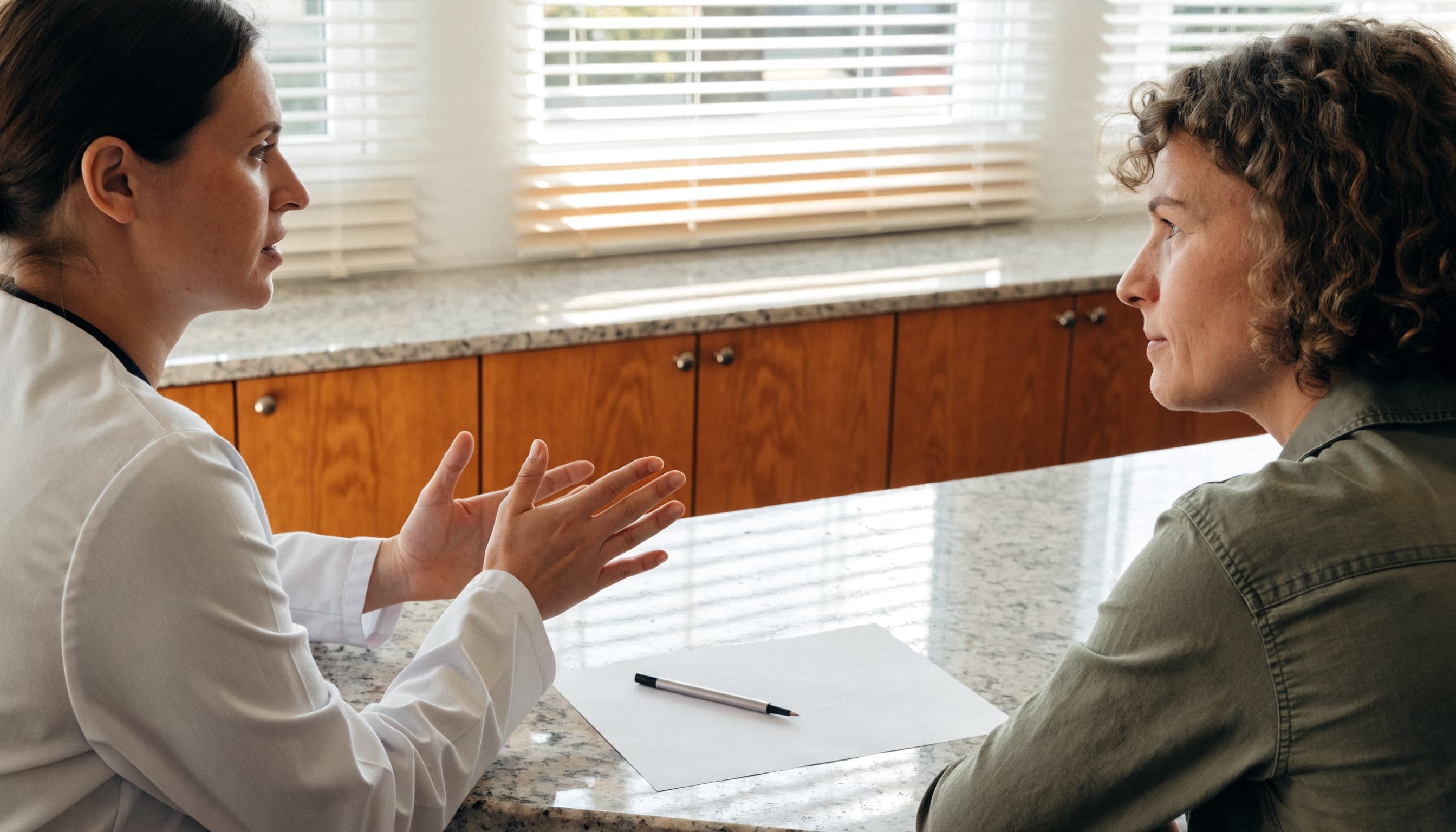 A clinician in a white coat explaining coverage to a patient across the office counter, a blank sheet of paper and a pen between them