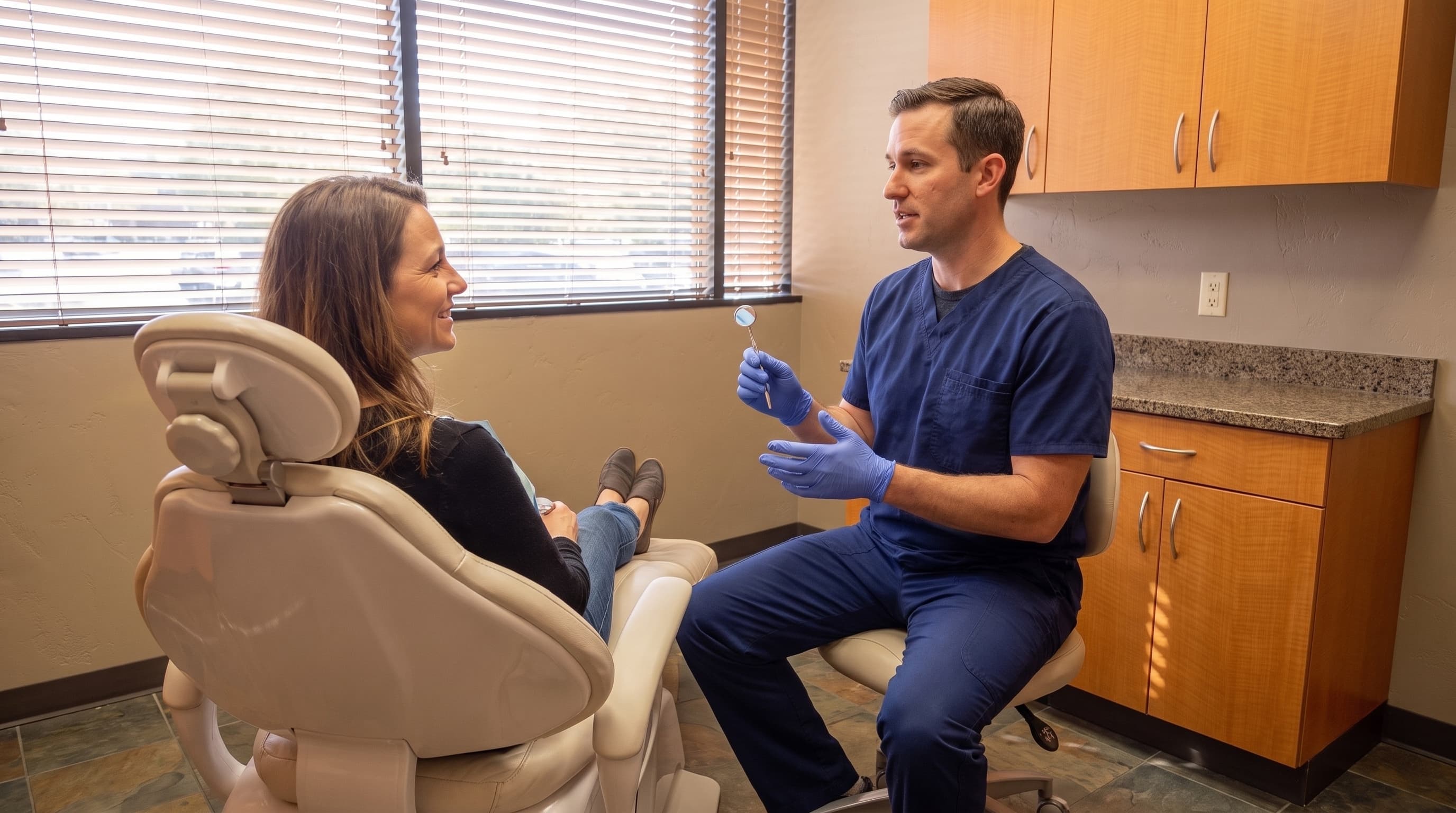 A clinician seated on a stool with a small dental mirror in hand, talking with a smiling patient reclined in the treatment chair