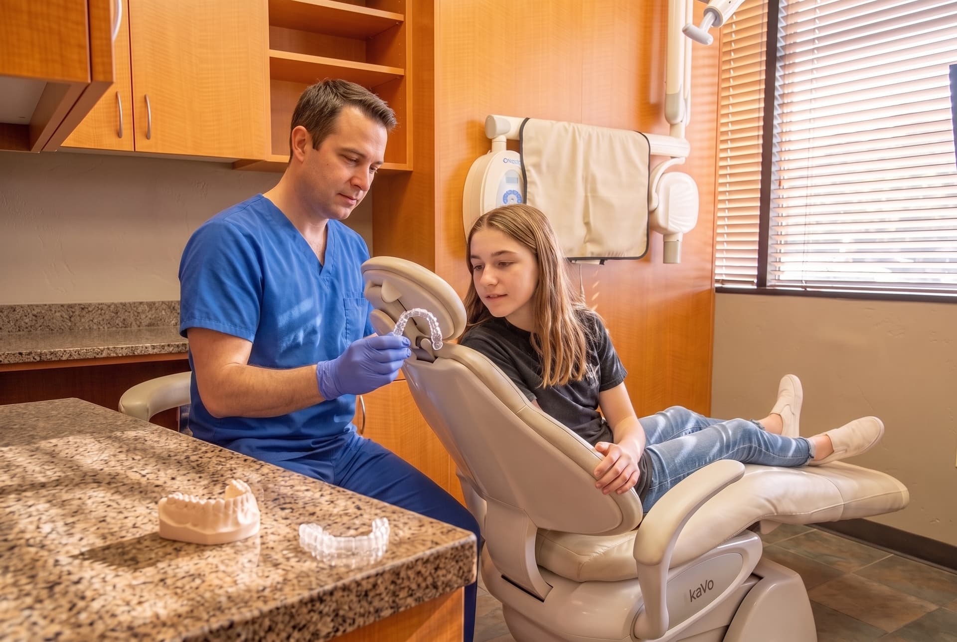 A clinician holding up a clear aligner tray for a teenage patient in the treatment chair, a plaster model on the counter