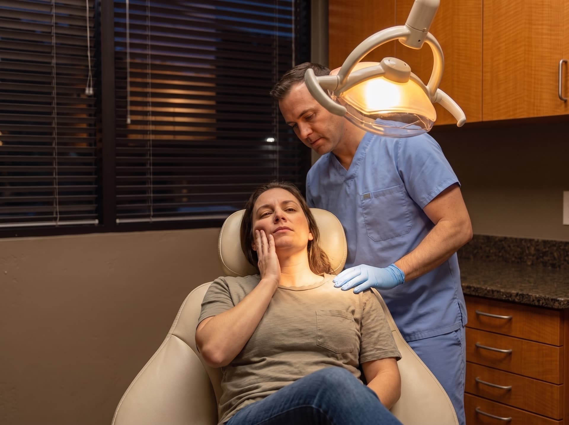 A patient in the treatment chair with a hand to the cheek while a clinician leans in under the overhead light
